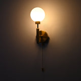 Raw brass wall sconce with a round frosted glass globe shade, illuminated, featuring a pull chain switch against a dark wall.