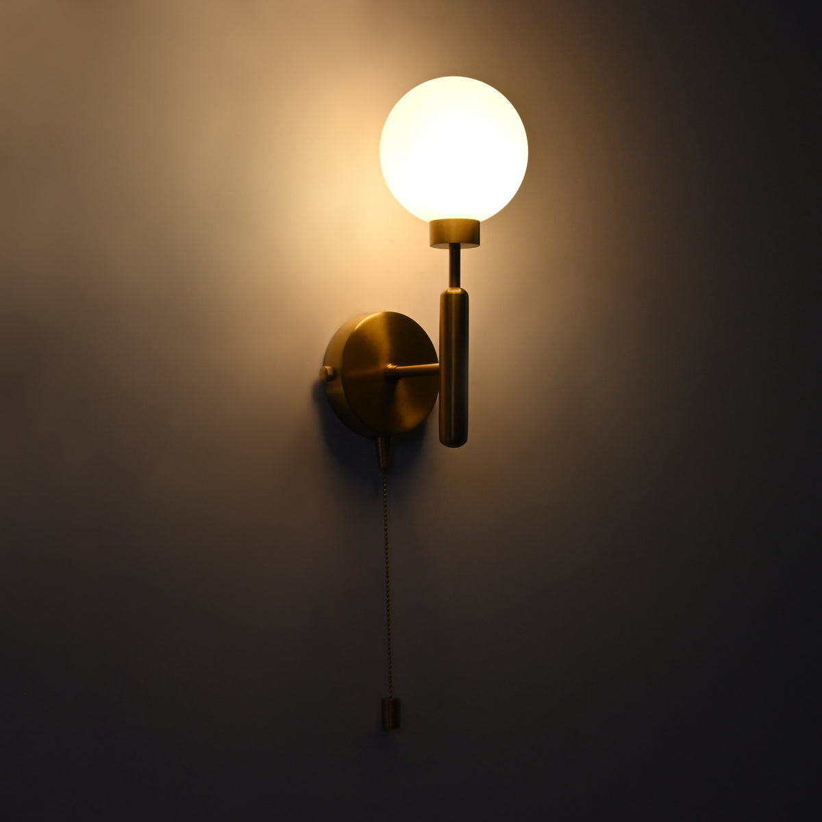 Raw brass wall sconce with round frosted glass globe illuminated, pull chain switch, mounted on dark wall.