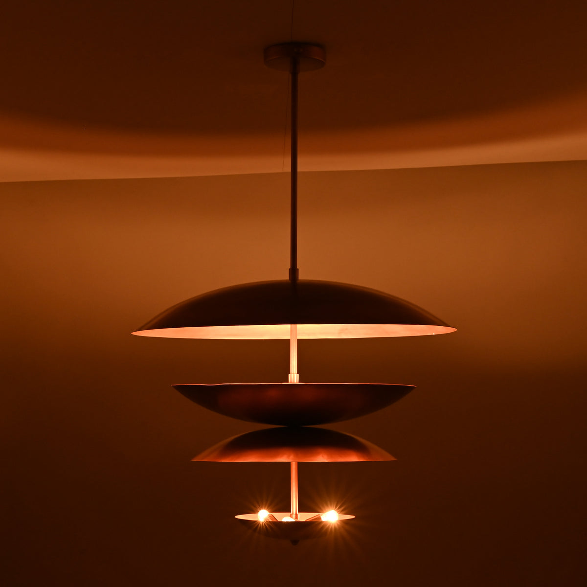 Lit view of the floating disc-style brass chandelier with stacked bowl elements and warm glowing indirect lights.