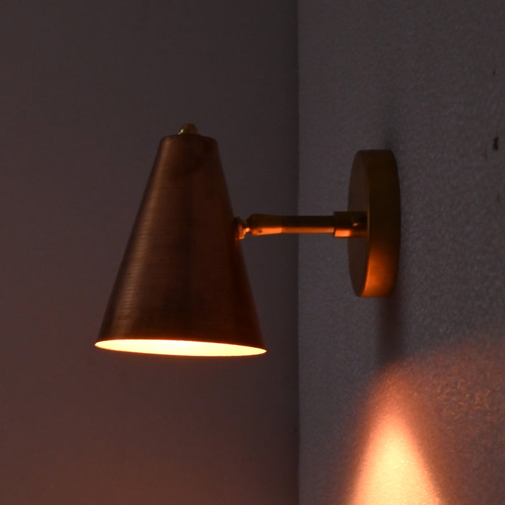 Industrial brass wall sconce with copper cone shade, glowing in dark interior for accent lighting.