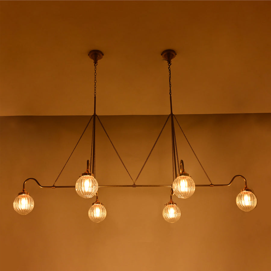 Linear 6-light ribbed glass globe chandelier shown in a warm bedroom interior with brass frame and ambient ceiling lighting