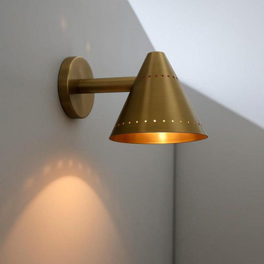 Minimalist brass cone wall sconce casting warm ambient light, highlighting perforated detailing and soft indirect illumination.