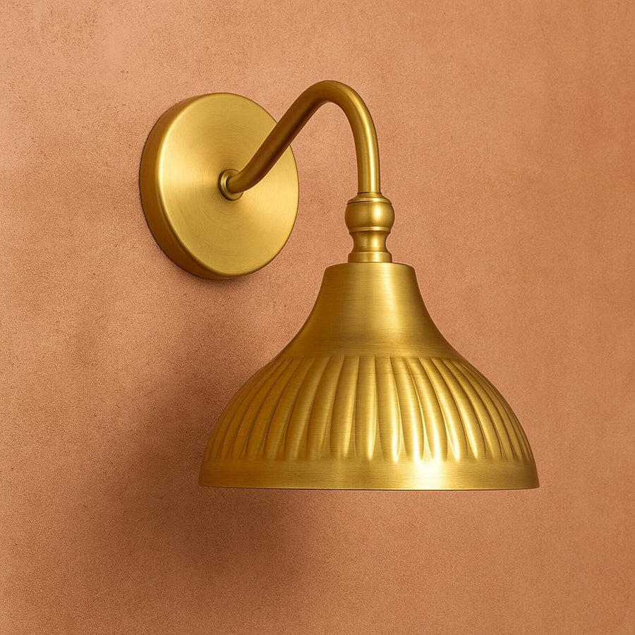 Modern brushed brass fluted dome wall sconce with curved arm mounted on warm peach textured wall background.