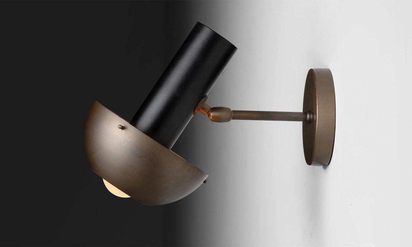 Modern brass wall lamp with sculptural bowl shade and cylindrical form, casting warm ambient light in a refined industrial design.