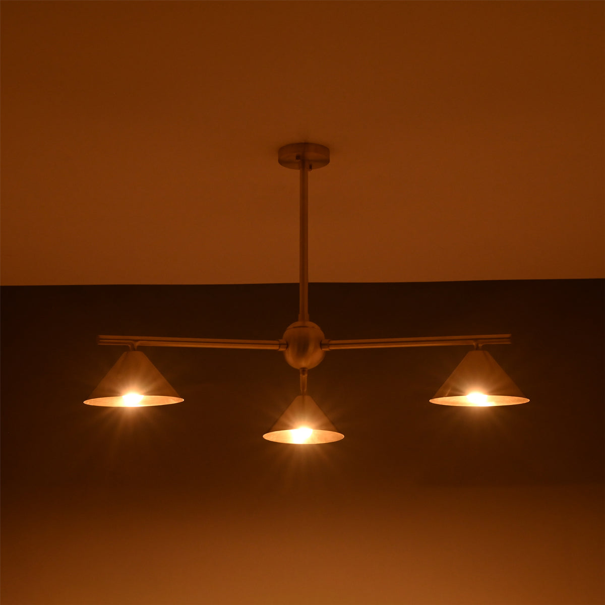 Handcrafted brass cone chandelier with three lights glowing softly in ambient warm lighting.