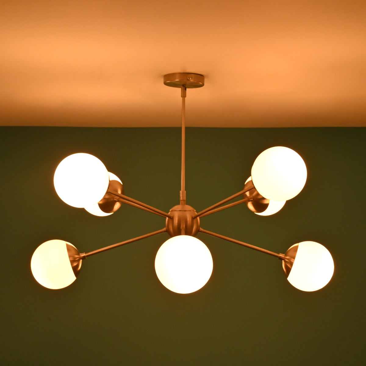 Modern brushed nickel chandelier with warm glowing globes illuminating a green wall backdrop.