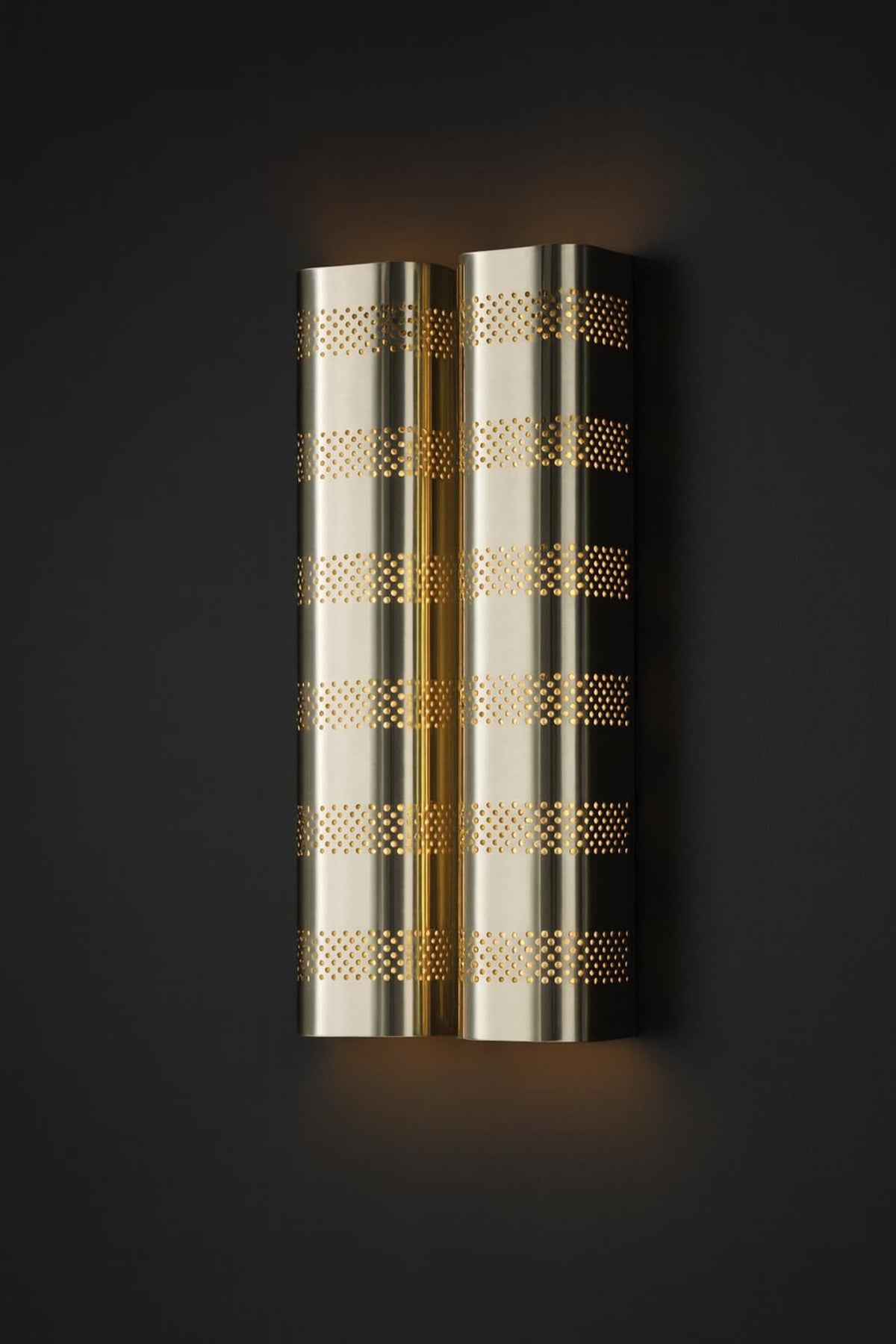 Polished brass perforated wall sconce with warm indirect light displayed against dark background