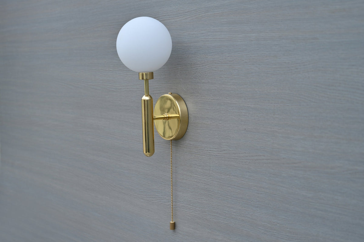Angled view of minimalist polished brass wall sconce with opal glass shade and chain pull detail
