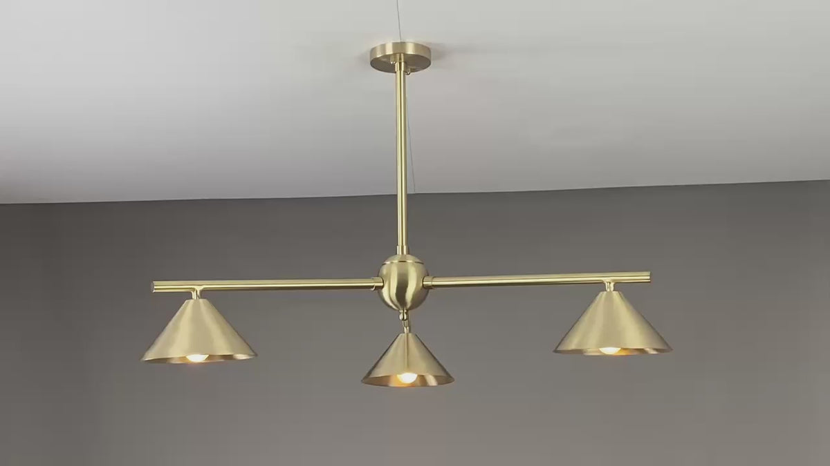 Warm-lit modern brass chandelier with three cone-shaped shades, creating cozy ambient lighting in a contemporary living room setting