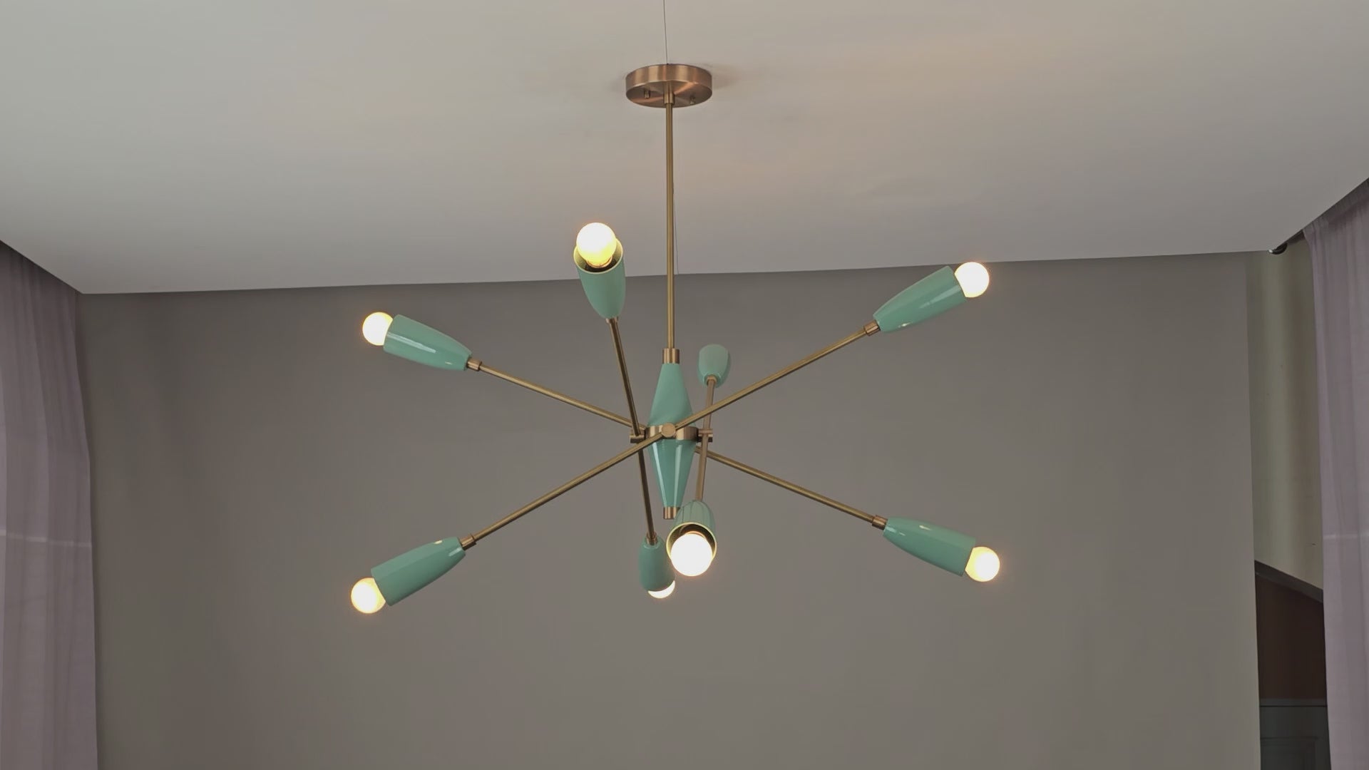 Mid-century modern Sputnik chandelier featuring eight warm-glowing bulbs with teal cone-shaped shades and brass arms, viewed from below against a ceiling with a soft amber lighting ambiance