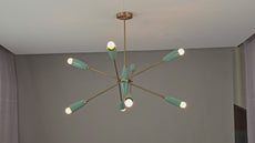 Mid-century modern Sputnik chandelier featuring eight warm-glowing bulbs with teal cone-shaped shades and brass arms, viewed from below against a ceiling with a soft amber lighting ambiance
