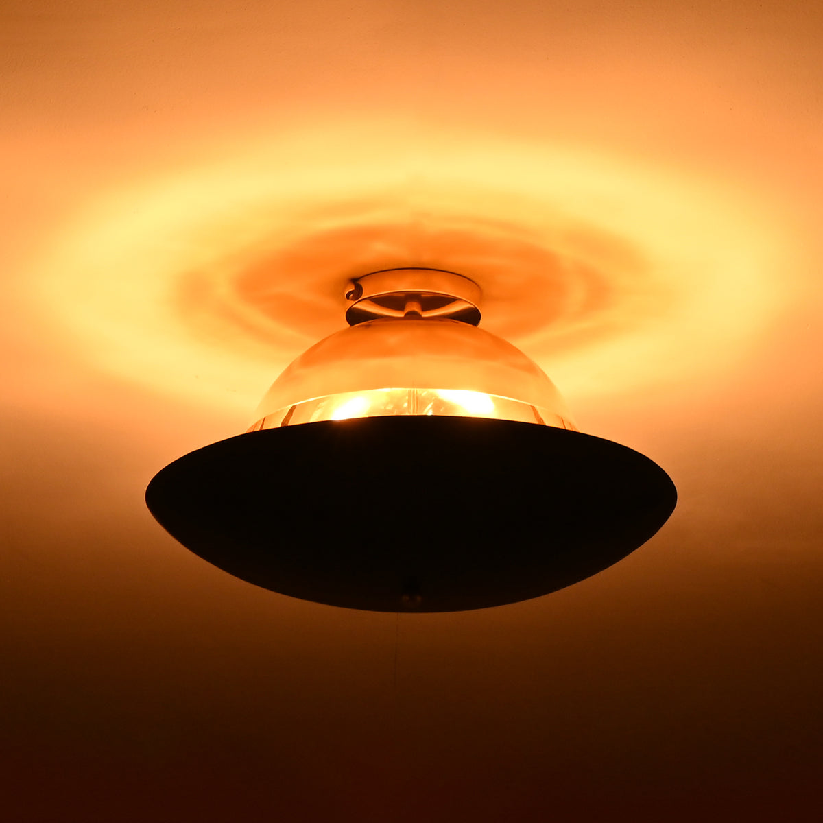 Two-tone brass ceiling light illuminated in low light, revealing polished brass upper dome and blackened disc with warm ambient glow.