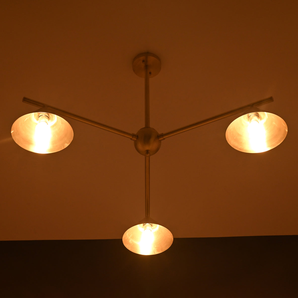 Brushed brass 3-light chandelier illuminated in warm ambient lighting, showcasing cone shades in a dark room.